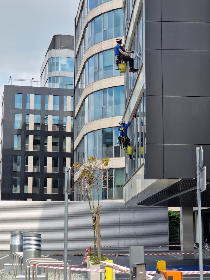 Two workers clean windows on a tall office building in a city setting.